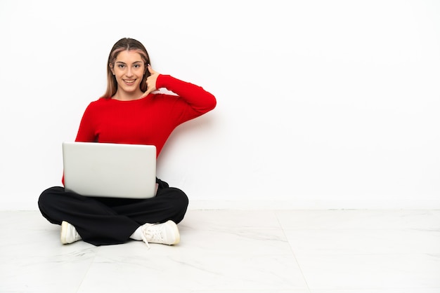 Jeune femme caucasienne avec un ordinateur portable assis sur le sol en faisant un geste de téléphone. Rappelez-moi signe
