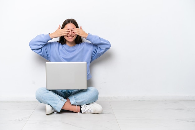 Jeune femme caucasienne avec ordinateur portable assis sur le sol isolé sur fond blanc couvrant les yeux par les mains