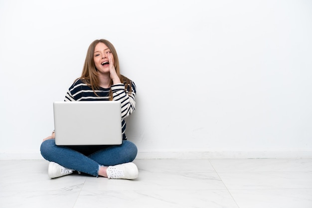 Jeune femme caucasienne avec un ordinateur portable assis sur le sol isolé sur fond blanc criant avec la bouche grande ouverte