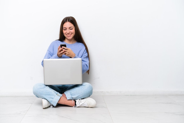 Jeune femme caucasienne avec ordinateur portable assis sur le sol isolé sur fond blanc envoyant un message avec le mobile