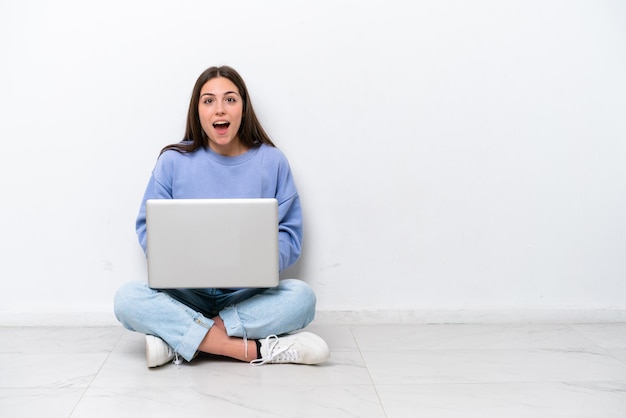 Jeune femme caucasienne avec ordinateur portable assis sur le sol isolé sur fond blanc avec une expression faciale surprise