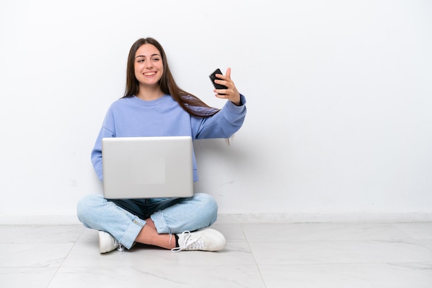 Jeune femme caucasienne avec ordinateur portable assis sur le sol isolé sur fond blanc faisant un selfie