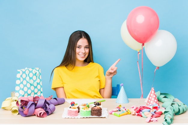 Jeune femme caucasienne organisant un anniversaire souriant et pointant de côté, montrant quelque chose à l'espace vide.