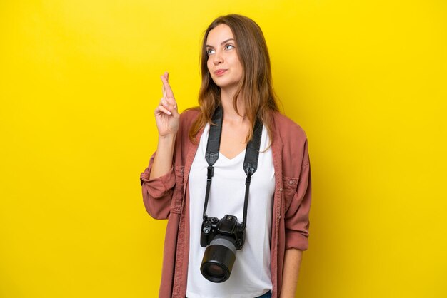 Jeune femme caucasienne photographe isolée sur fond jaune avec les doigts croisés et souhaitant le meilleur