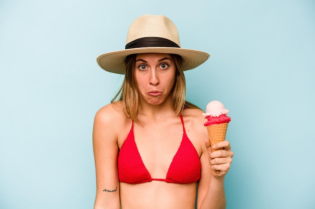 Jeune femme caucasienne portant un bikini et tenant une glace isolée sur fond bleu hausse les épaules et ouvre les yeux confus