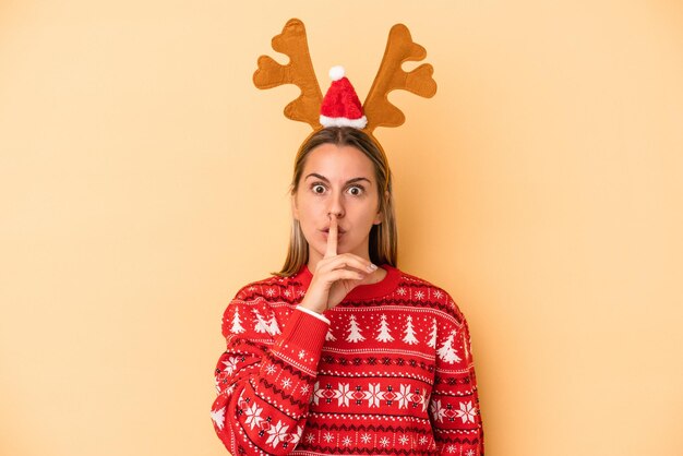 Jeune femme caucasienne portant un chapeau de renne de noël isolé sur fond jaune gardant un secret ou demandant le silence.