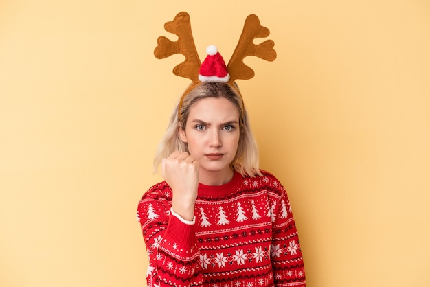 Jeune femme caucasienne portant un chapeau de renne de Noël isolé sur fond jaune montrant le poing à la caméra, expression faciale agressive.