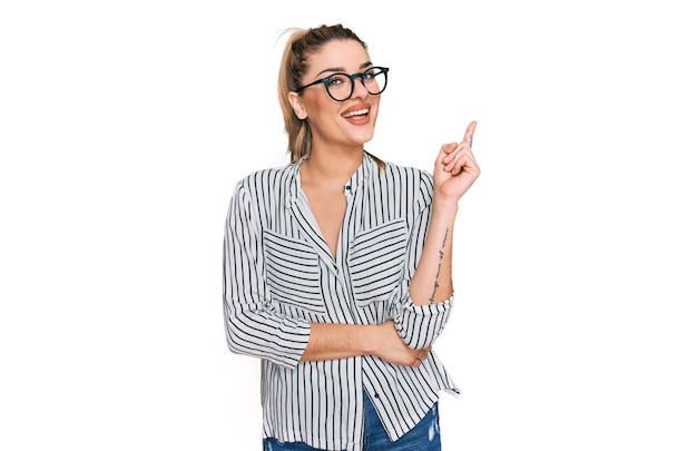 Jeune femme caucasienne portant une chemise d'affaires et des lunettes avec un grand sourire sur le visage, pointant avec la main et le doigt sur le côté en regardant la caméra.