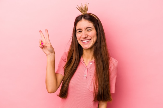 Jeune femme caucasienne portant une couronne isolée sur fond rose joyeuse et insouciante montrant un symbole de paix avec les doigts.