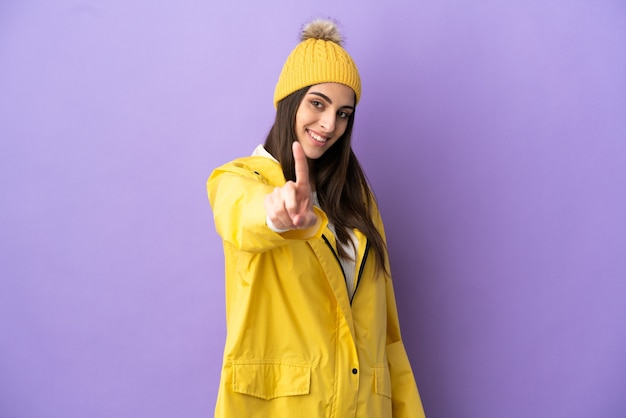 Jeune femme caucasienne portant un manteau imperméable isolé sur fond violet montrant et levant un doigt