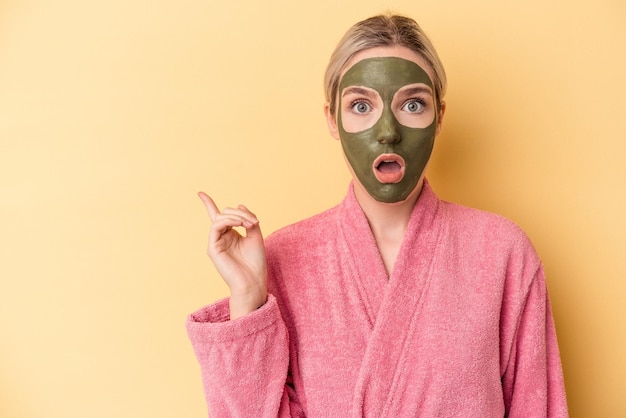 Jeune femme caucasienne portant un masque facial isolé sur fond jaune pointant vers le côté