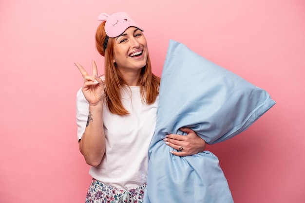 Jeune femme caucasienne portant un pyjama tenant un oreiller isolé sur fond rose joyeux et insouciant montrant un symbole de paix avec les doigts.