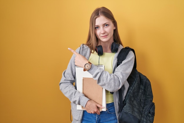 Jeune femme caucasienne portant un sac à dos étudiant et tenant des livres pointant avec le doigt de la main sur le côté montrant la publicité visage sérieux et calme