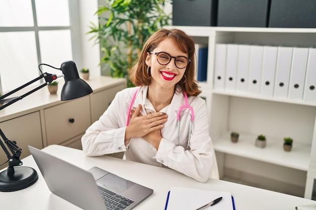 Jeune femme caucasienne portant un uniforme de médecin avec les mains sur le cœur à la clinique