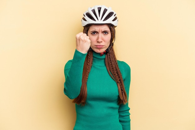 Jeune femme caucasienne portant un vélo casque isolé sur fond jaune montrant le poing à la caméra, expression faciale agressive.
