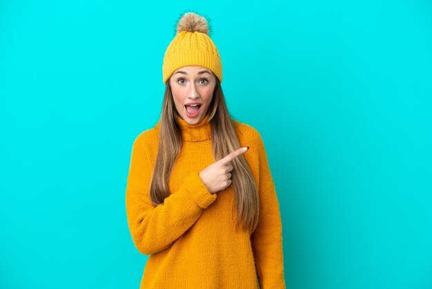 Jeune femme caucasienne portant une veste d'hiver isolée sur fond bleu surpris et pointant du côté