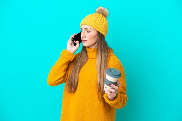 Jeune femme caucasienne portant une veste d'hiver isolée sur fond bleu tenant du café à emporter et un mobile