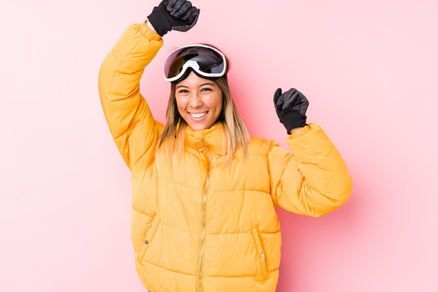 Jeune femme caucasienne portant un vêtement de ski dans un mur rose célébrant une journée spéciale, saute et lève les bras avec énergie.