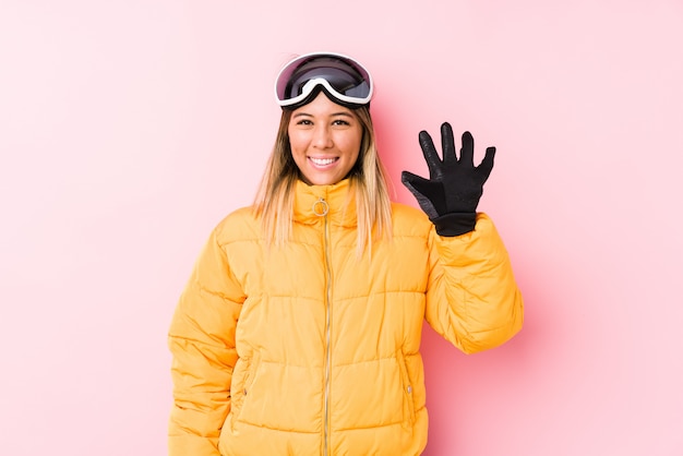 Jeune femme caucasienne portant un vêtement de ski dans un mur rose souriant joyeux montrant le numéro cinq avec les doigts.