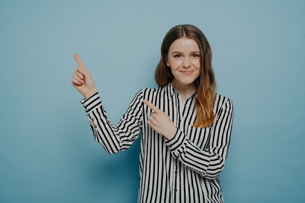 Jeune femme caucasienne positive vêtue d'une chemise rayée pointant vers le coin supérieur gauche avec les index et souriant à la caméra, isolée sur fond bleu. Publicité et promotion