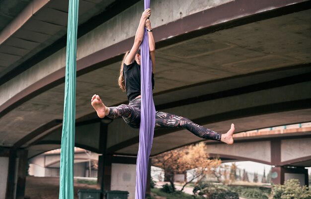 Jeune femme caucasienne pratiquant la performance de la soie aérienne accrochée à un pont dans une ville de parc, entourée par la nature.