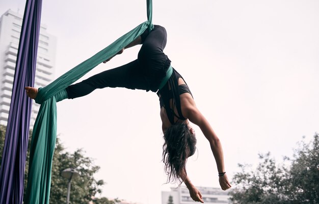 Jeune femme caucasienne pratiquant la performance de la soie aérienne accrochée à un pont dans une ville de parc, entourée par la nature.