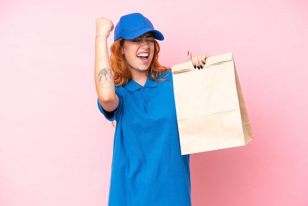 Jeune femme caucasienne prenant un sac de plats à emporter isolé sur fond rose célébrant une victoire