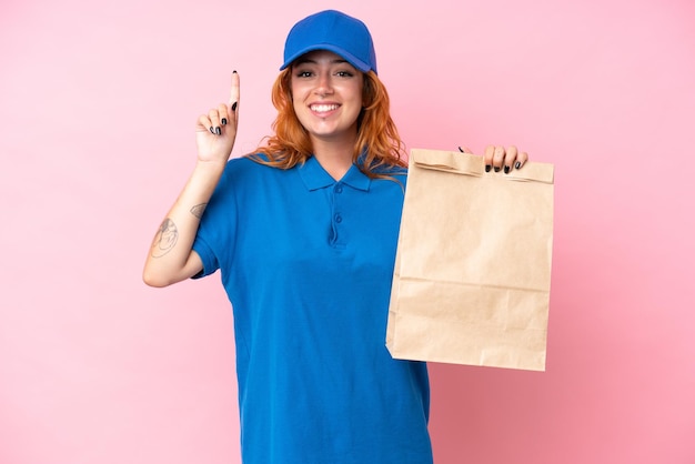 Jeune femme caucasienne prenant un sac de plats à emporter isolé sur fond rose pointant vers le haut une excellente idée