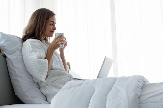 Jeune femme caucasienne en pyjama blanc avec du blanc tenant une tasse de café et une boisson et trouver des informations à partir de l'ordinateur portable sur un lit dans une chambre à la maison. Femme buvant du café et travaillant sur un ordinateur portable.