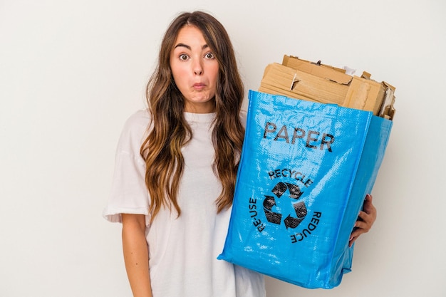 Jeune femme caucasienne recyclant du papier isolé sur fond blanc hausse les épaules et ouvre les yeux confus.