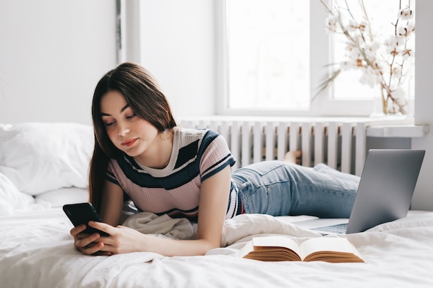 Jeune femme caucasienne reposant sur un lit à la maison et utilisant un ordinateur portable et un smartphone.