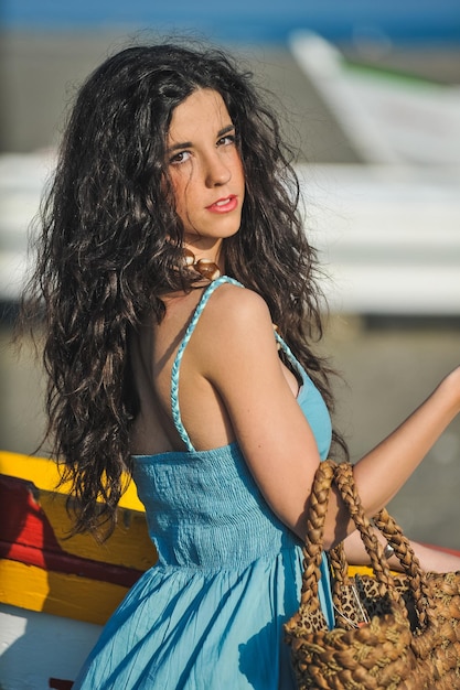 Jeune femme caucasienne avec robe bleu clair et sac à main à côté d'un bateau dans le sable sur la plage