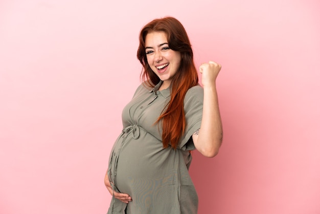 Jeune femme caucasienne rousse isolée sur fond rose enceinte et fête une victoire