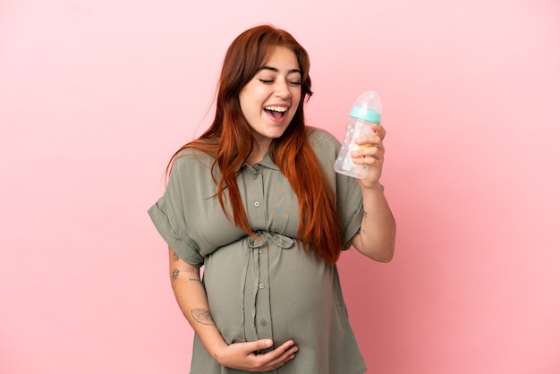 Jeune femme caucasienne rousse isolée sur fond rose enceinte et tenant un biberon avec une expression surprise