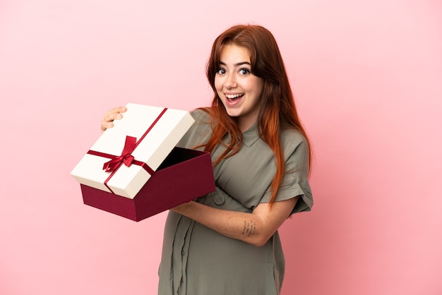Jeune femme caucasienne rousse isolée sur fond rose enceinte et tenant un cadeau avec une expression surprise