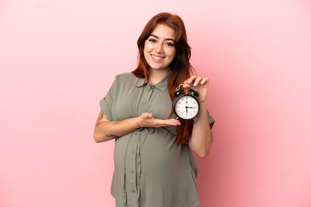 Jeune femme caucasienne rousse isolée sur fond rose enceinte et tenant l'horloge