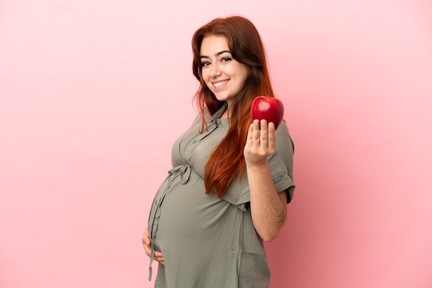 Jeune femme caucasienne rousse isolée sur fond rose enceinte et tenant une pomme