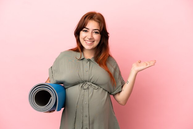 Jeune femme caucasienne rousse isolée sur fond rose enceinte et tenant un tapis tout en présentant quelque chose