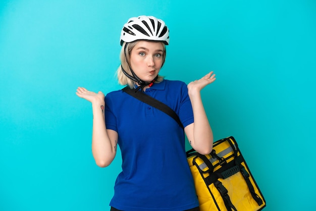 Jeune femme caucasienne avec sac à dos thermique isolé sur fond bleu ayant des doutes tout en levant les mains