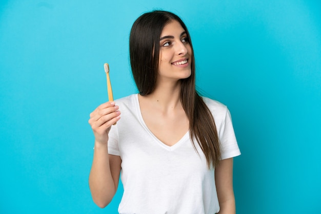 Jeune femme caucasienne se brosser les dents isolées sur fond bleu en levant tout en souriant