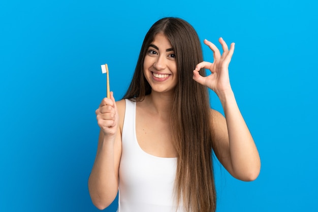 Jeune femme caucasienne se brosser les dents isolées sur fond bleu montrant un signe ok avec les doigts