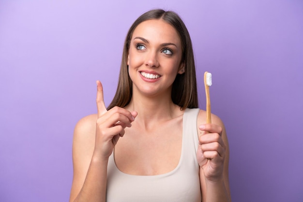 Jeune femme caucasienne se brosser les dents isolées sur fond violet dans l'intention de réaliser la solution tout en levant un doigt