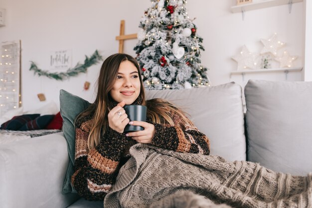 Jeune femme caucasienne se détendre sur un canapé à la maison et tenant une tasse avec une boisson chaude en vacances de Noël.