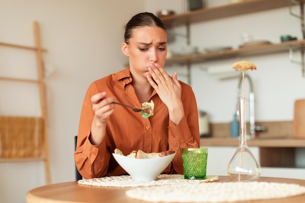 Jeune femme caucasienne se sentant nauséeuse en mangeant de la salade césar en regardant l'assiette et en fermant la bouche