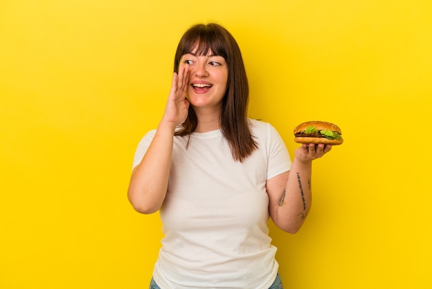 Jeune femme caucasienne sinueuse tenant un hamburger isolé sur fond jaune criant et tenant la paume près de la bouche ouverte.