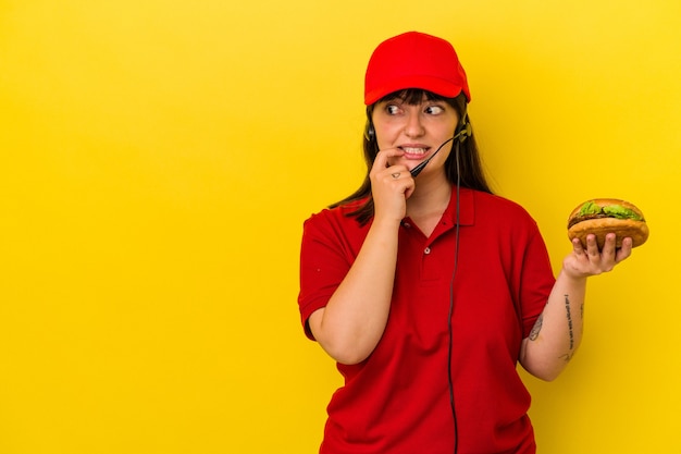 Jeune femme caucasienne sinueuse tenant un hamburger isolé sur fond jaune détendue en pensant à quelque chose en regardant un espace de copie.