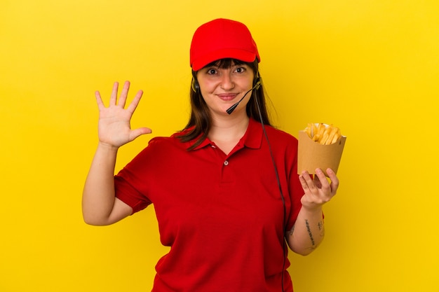 Jeune femme caucasienne sinueuse travailleur de restauration rapide tenant des frites isolées sur fond bleu souriant joyeux montrant le numéro cinq avec les doigts.