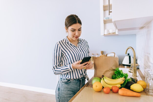 Jeune femme caucasienne souriante utilise le smartphone dans la cuisine moderne, sac avec des légumes frais sur la table. Achat en ligne de nourriture et d'épicerie.