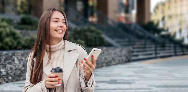 Une jeune femme caucasienne souriante utilise un téléphone portable et s'assoit sur fond d'un immeuble de bureaux