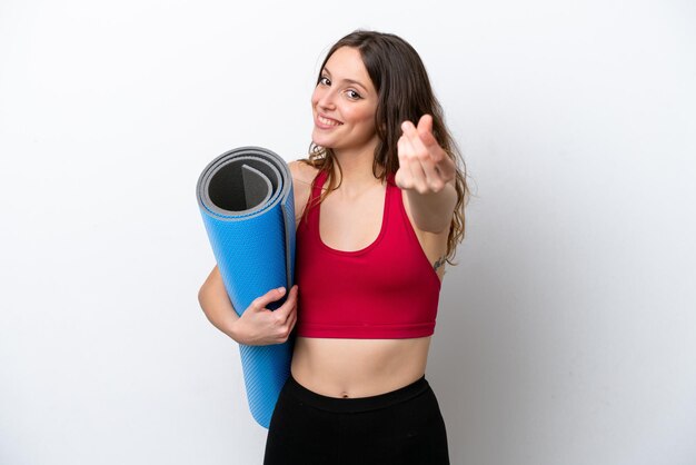 Jeune femme caucasienne sportive allant à des cours de yoga tout en tenant un tapis isolé sur fond blanc faisant un geste d'argent